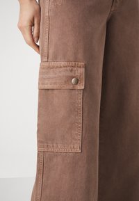 Pantalon cargo marron avec une poche latérale munie d'un bouton-pression métallique, confectionné en tissu texturé, avec une coupe à jambes larges et des détails de couture minimaux.