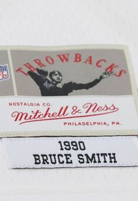 Étiquette en tissu blanc avec surface texturée ; comporte du texte rouge et noir : "THROWBACKS," "Mitchell & Ness," et "1990 BRUCE SMITH."