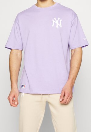 Mężczyzna noszący jasnofioletową koszulkę z krótkim rękawem z białym logo New York Yankees na piersi i beżowe spodnie.