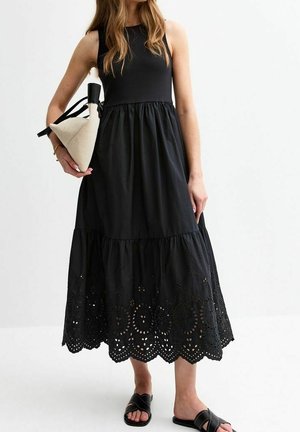 Kobieta w czarnej sukience bez rękawów z ażurowym wykończeniem, trzymająca beżową trójkątną torebkę, ubrana w czarne klapki, stoi na białym tle.