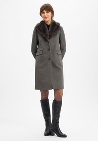 Grijze wollen jas met een pluche donkere bontkraag, voorzien van twee voorzakken, knoopsluiting en een rechte silhout. Een paar zwarte knie-hoge laarzen.