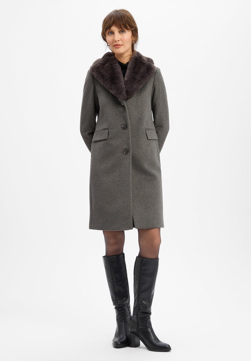 Grijze wollen jas met een pluche donkere bontkraag, voorzien van twee voorzakken, knoopsluiting en een rechte silhout. Een paar zwarte knie-hoge laarzen.