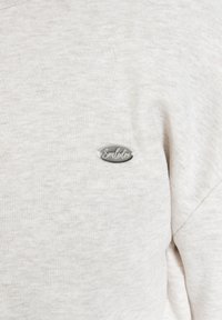 Felpa grigia chiara con una texture morbida. Presenta un piccolo logo ovale argentato sul petto con la scritta "Smulodex" incisa. Design semplice.