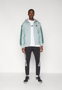 Φωτεινό γαλάζιο φούτερ με φερμουάρ και πράσινες ρίγες, λευκό μπλουζάκι, μαύρο στενό παντελόνι jogging με λευκές ρίγες και μαύρα αθλητικά παπούτσια.