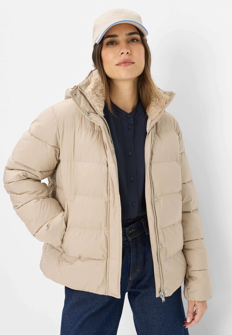 Beige pufferjakke med en struktureret imiteret pelskrave; lynlåslukning; sidelommer; båret over en mørk skjorte med knaplukning og blå jeans.