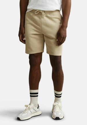Mann trägt beige Kordelzug-Shorts, weiß-schwarz gestreifte Socken und weiße Sneakers, steht vor einem einfarbigen Hintergrund.