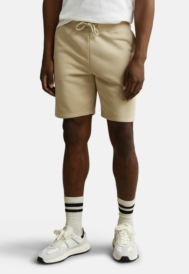 Homme portant un short beige à cordon, des chaussettes rayées blanches et noires, et des baskets blanches, debout devant un fond uni.