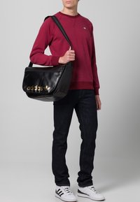 Svart syntetisk budväska med guld "adidas"-logotyp, justerbar rem och blixtlåsficka. Modellen bär en vinröd sweatshirt och mörka jeans.