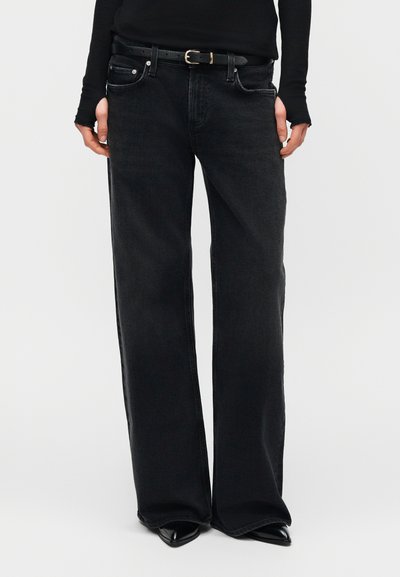 Μαύρα τζιν φαρδιάς γραμμής, φορεμένα με μαύρα παπούτσια μυτερά και μαύρο μακρυμάνικο τοπ, τα χέρια χαλαρά στα πλάγια.