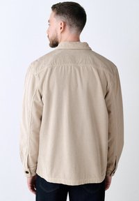 Man with short hair and beard wearing a beige corduroy long-sleeve shirt, facing away against a plain white background.