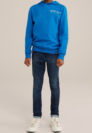 Dziecko w jasnoniebieskiej bluzie z kapturem z napisem, ciemnoniebieskich dżinsach i białych tenisówkach, stojące na neutralnym tle.