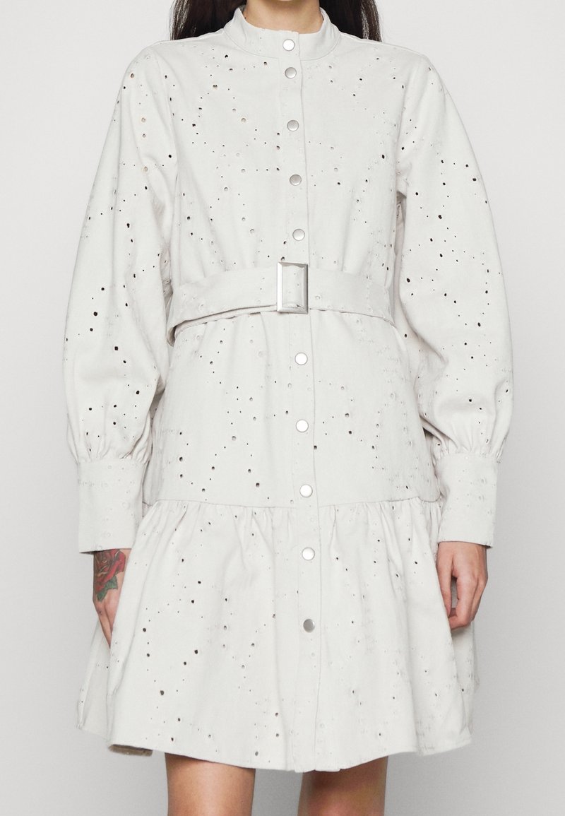 Robe en coton gris clair avec un col arrondi, des manches longues et une patte de boutonnage à l'avant, ceinturée à la taille et ornée de détails perforés sur toute la surface.
