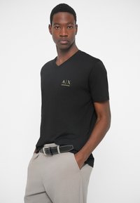 Μαύρο μπλουζάκι v-neck από απαλό ύφασμα, με ένα μικρό χρυσό λογότυπο στο στήθος. Συνδυάζεται με ανοιχτό γκρι παντελόνι και μαύρη ζώνη.