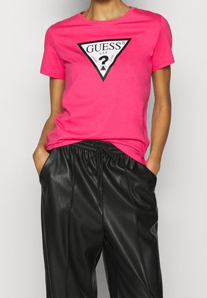 Persoon draagt een felroze korte mouwen GUESS T-shirt met zwart driehoeklogo en zwarte nepleren broek met trekkoord, handen in de zakken.