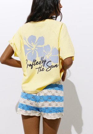 Femme portant un t-shirt jaune à manches courtes avec un imprimé floral bleu et le texte « Crafted by the Sun », portant un short crocheté bleu et blanc à rayures.