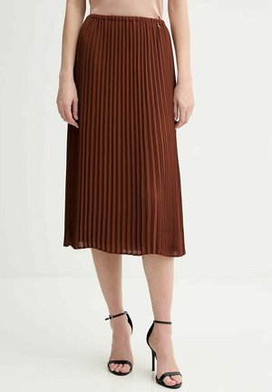 Donna che indossa una gonna midi plissettata marrone e sandali neri con tacco alto e cinturino alla caviglia, in piedi contro uno sfondo semplice.