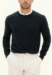 Marinblå tröja med rund halsringning och långa ärmar, tillverkad av ett mjukt material. Bärs över en vit skjorta med krage, ihop med creamfärgade byxor.