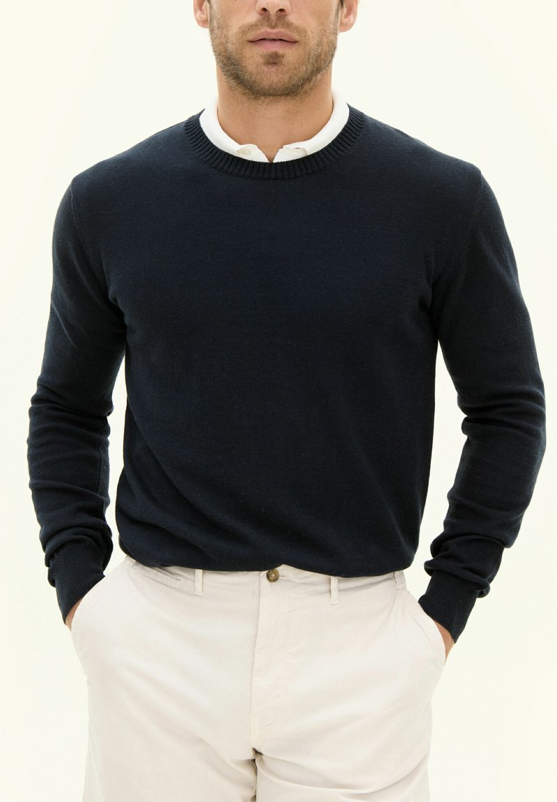 Marinblå tröja med rund halsringning och långa ärmar, tillverkad av ett mjukt material. Bärs över en vit skjorta med krage, ihop med creamfärgade byxor.