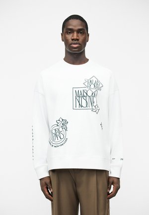 Homme noir portant un sweat-shirt blanc avec des motifs floraux et le texte « Maison Kitsuné » et « Tokyo Paris », associé à un pantalon marron.