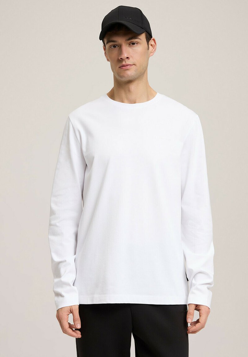 Biała koszula z długim rękawem i okrągłym dekoltem, uszyta z gładkiego materiału, noszona z czarnymi spodniami. Czarna czapka dopełnia strój.