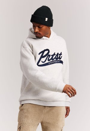 Man wearing black knit beanie, white "Prtst Sportswear" hoodie, and beige fleece pants standing against plain background looking down.