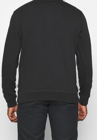 Sweat-shirt noir à manches longues, col rond, et poignets côtelés. Vue arrière montrant des détails de couture subtils et une étiquette logo à l'ourlet.