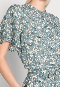 Blusa in tessuto turchese a fantasia floreale con maniche corte a sbuffo, scollo rotondo e bottoni sul davanti; dettaglio della vita elastica visibile.