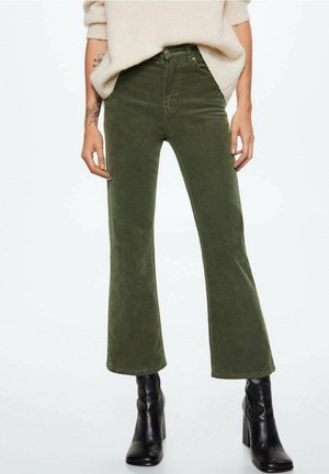 Personne portant un pantalon en velours côtelé vert taille haute, un pull beige et des bottines noires à talons, debout avec les bras détendus le long du corps.