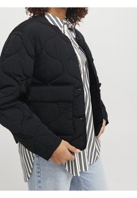 Předpřený černý kabát s texturovaným povrchem, s přední kapsou a knoflíkovým zapínáním, nošený přes pruhovanou košili a džínami.