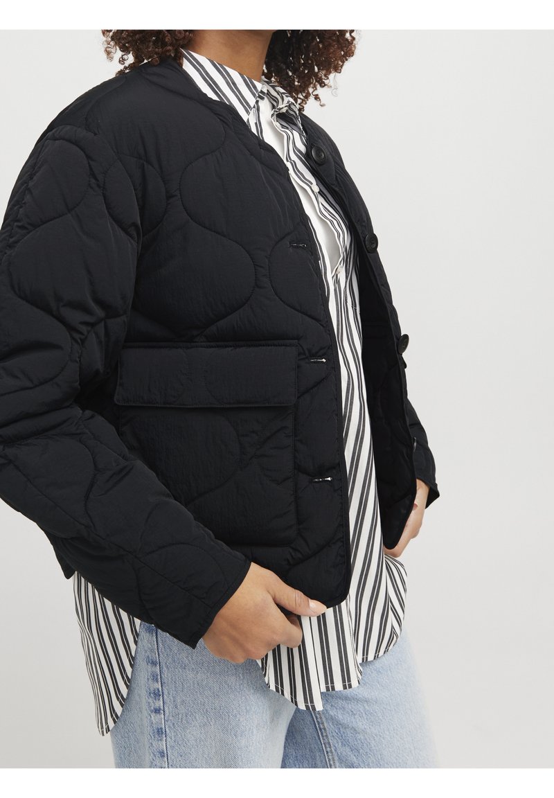 Předpřený černý kabát s texturovaným povrchem, s přední kapsou a knoflíkovým zapínáním, nošený přes pruhovanou košili a džínami.