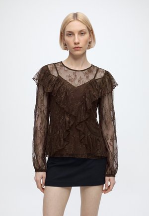Kvinde med kort blondt hår iført en gennemsigtig brun blondebluse med flæser og et sort skørt, stående mod en ensfarvet baggrund.