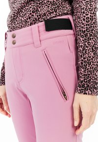 Ružové lyžiarske nohavice s hladkou textúrou, obsahujúce zipsovú kapsu, dvojité kovové gombíky a čierny suchý zips ako akcent na páse.