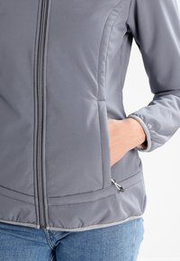 Veste softshell grise avec fermeture éclair à l'avant, poches latérales et poignets élastiques. Présente une texture lisse et élégante avec une bordure contrastante au bas.