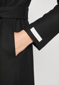 Zwarte wollen jas met een ceintuur in de taille, met een zijzak en een label op de manchette. De textuur is glad en het ontwerp is op maat gemaakt.