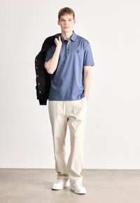 Modré polokošeľa s tromi gombíkmi a logom na hrudi, v kombinácii s béžovými nohavicami a bielymi teniskami, čierna bunda prehodená cez plece.