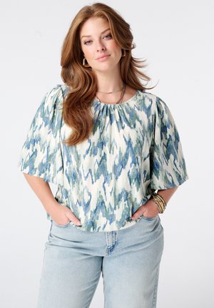 Vrouw met lang bruin haar, een blauw-groen dessin blouse en lichtblauwe spijkerbroek, handen in de zakken, kijkend naar voren tegen een witte achtergrond.