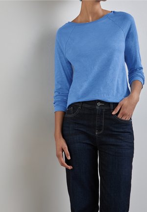 Person trägt ein hellblaues Langarmhemd und dunkelblaue Jeans, steht mit einer Hand in der Tasche vor einem schlichten Hintergrund.