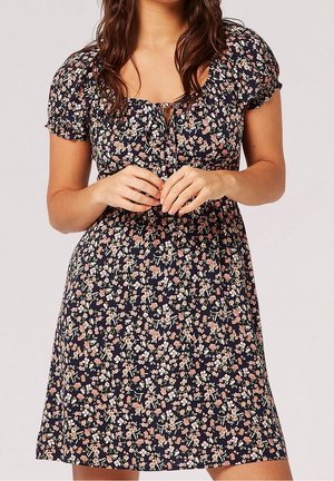 Kobieta ubrana w granatową sukienkę z krótkimi bufkami na rękawach oraz różowo-białym kwiatowym wzorem, delikatnie splecione dłonie z przodu.