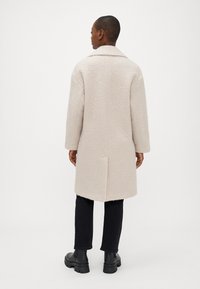 Ανοιχτό καφέ υφασμάτινο παλτό από μάλλινο ύφασμα με ανάγλυφη υφή, με κομμένο γιακά, μακριά μανίκια και ίσια σιλουέτα, διαθέτοντας άνοιγμα στη πλάτη για εύκολη κίνηση.