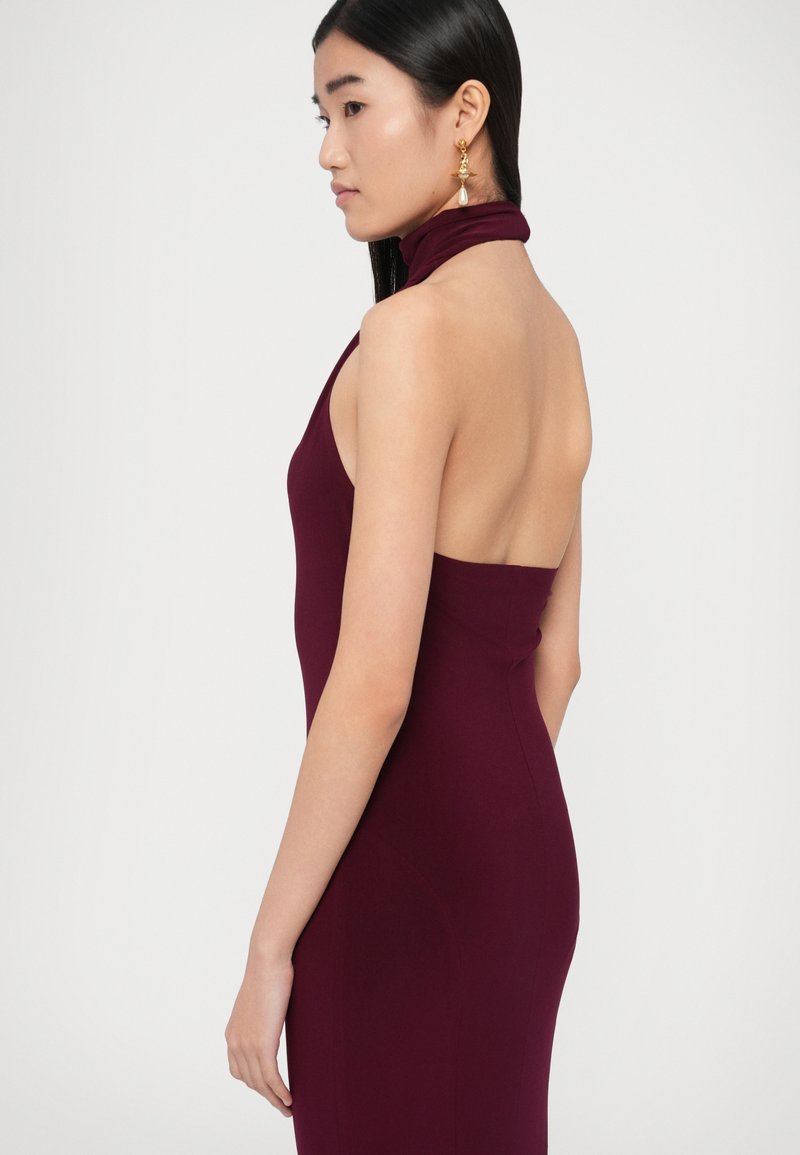 Robe sans manches bordeaux avec un col montant et un dos nu. Tissu lisse avec une silhouette moulante. Boucles d'oreilles pendantes dorées mises en valeur.