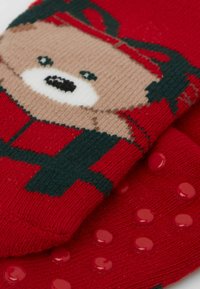 Calzini rossi con design di un orso dal viso beige, dettagli verdi e puntini antiscivolo rossi sulla suola per una miglior presa. Tessuto lavorato a maglia e materiale caldo.