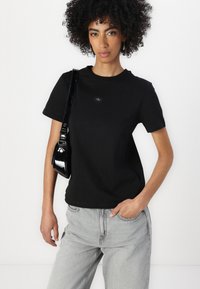 Svart bomull t-skjorte med korte ermer og rund hals, med en liten logo-patch på forsiden, parret med lysegrå jeans og en blank svart håndveske.