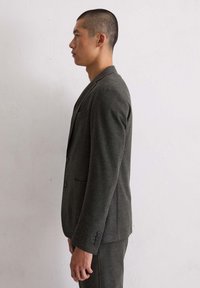 Man met kort haar in een donkergrijs pak staat in profiel tegen een effen witte muur.