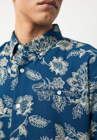 Camicia a maniche corte blu con motivo floreale bianco, colletto con bottoni e taschino sul petto con due bottoni sul davanti.