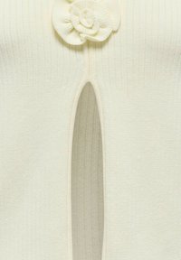 Gros plan sur un tissu tricoté côte crème avec une découpe en forme de trou de serrure et un détail de fleur décoratif en haut au centre.