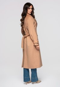 Beige villainen takki, jossa on vyötäröllä solmittava nauha, suuri kaulus ja pitkät hihat, yhdistettynä levenevillä lahkeilla varustettuihin sinisiin farkkuihin ja beigen värisiin korkokenkiin.