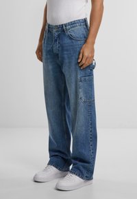 Pantalones vaqueros de mezclilla azul con un corte relajado, que presentan un gran bolsillo lateral, costuras en contraste y un lavado descolorido. Llevados con zapatillas blancas.
