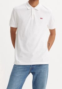 Bílá bavlněná polo košile s klasickým límečkem a třemi knoflíky. Na hrudi je malé červené logo Levi's. Je nošena s modrými džíny.