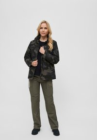 Bunda s camouflage vzorem v tmavě zelených a černých barvách, se zipem vpředu, límcem a kapsami. V kombinaci s olivovými cargo kalhotami a černými botami.