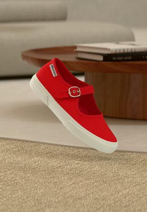 Scarpa Mary Jane in tela rossa con suola in gomma bianca e cinturino con fibbia argento, posizionata vicino a un tavolino in legno con libri impilati.